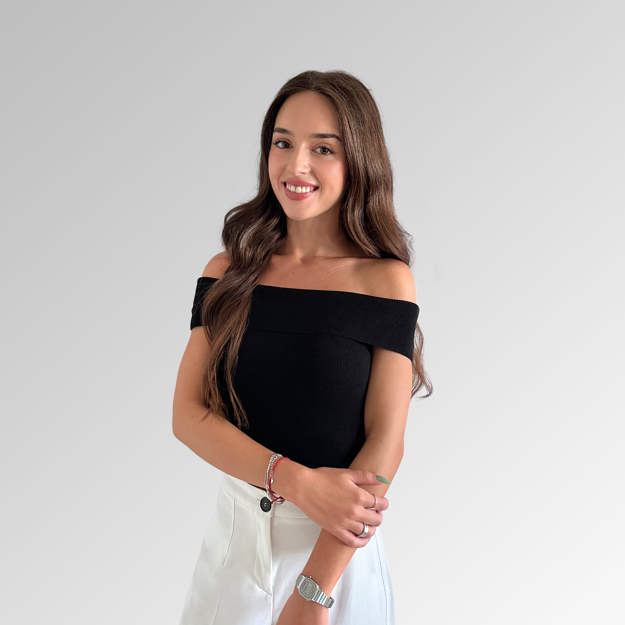 Confident young woman in off-the-shoulder top and high-waisted pants against a light gray background.