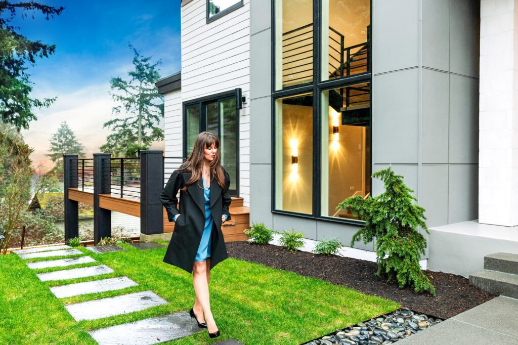 Elegant woman strolling towards a modern home surrounded by lush greenery at sunset.