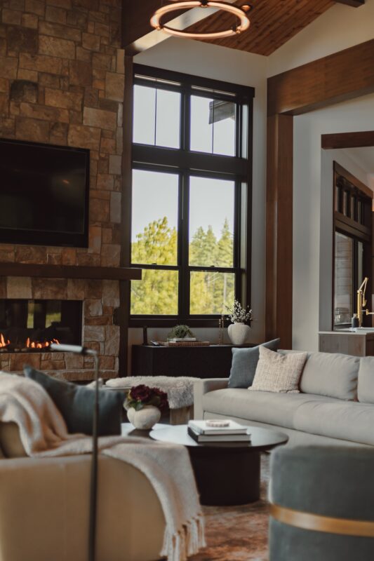 Corner view highlighting stone fireplace, black-framed windows, and natural light, styled with soft textiles and curated decorative details by Ariana Designs.