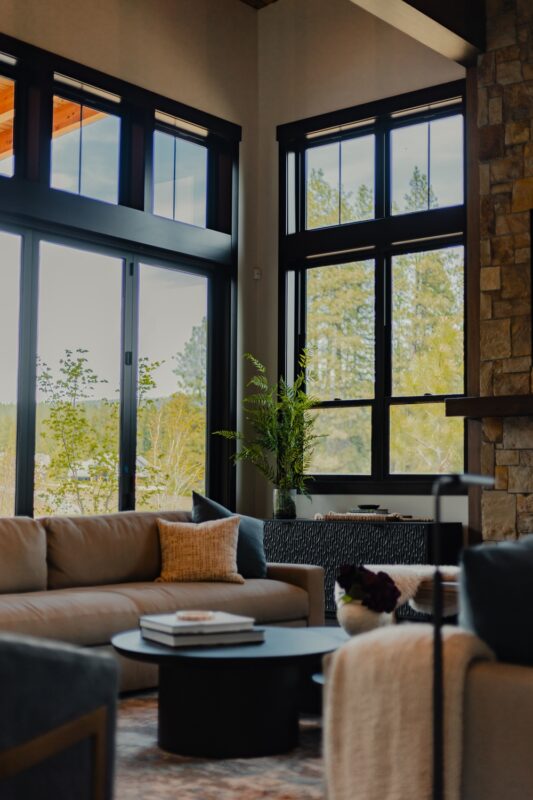 Stylish living space with circular black coffee table, soft throw blankets, and plush seating centered around panoramic windows and natural mountain light.