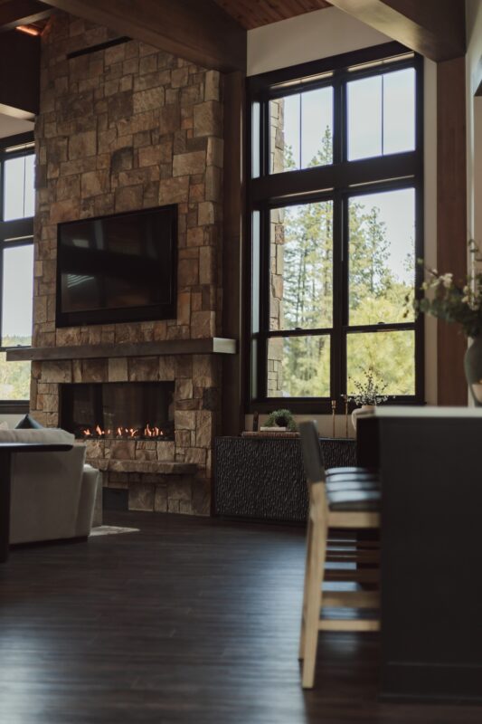 Side perspective of the family room showing the stone fireplace, soft seating, and natural light streaming through tall windows with dark wood framing.