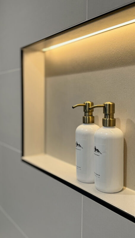 Minimalist shower niche with warm LED lighting highlighting white bottles with gold pump tops, accentuating the bathroom’s elegant spa aesthetic.