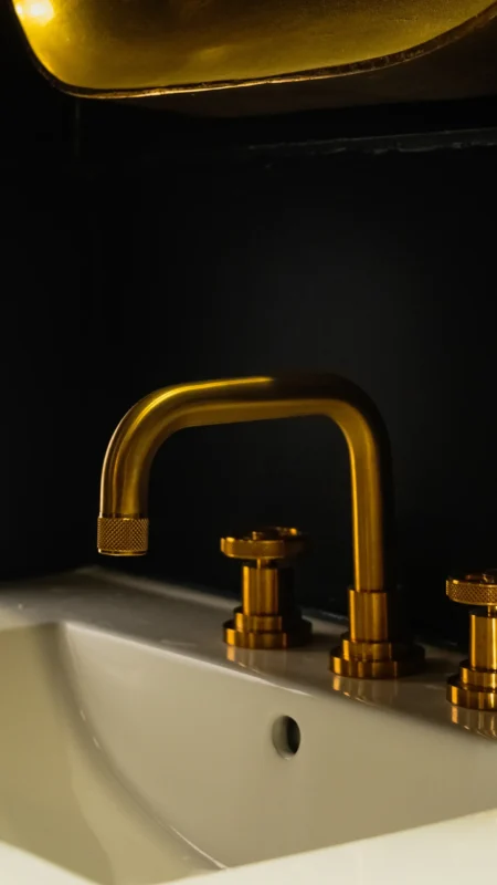 Close-up vertical shot of a brushed gold gooseneck faucet and matching knurled handles mounted on a white sink basin against a deep black wall.