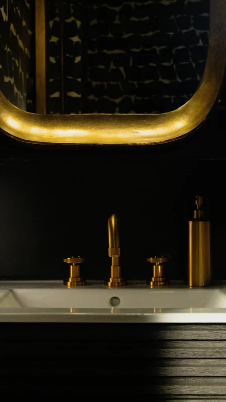 Close-up, horizontal view of gold bathroom faucet and handles on a white sink, set against a dark wall and gold-framed mirror.