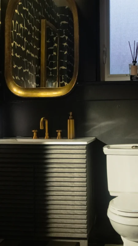 Vertical view of a dark, stylish powder room corner with a gold-framed mirror, fluted vanity, gold sink fixtures, and a white toilet bowl on the right.