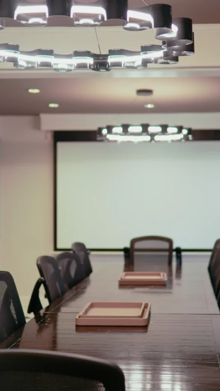 Close-up, low-angle shot of a conference table with dark wood finish and black mesh chairs in the foreground, with a large projection screen and a circular black chandelier visible above.