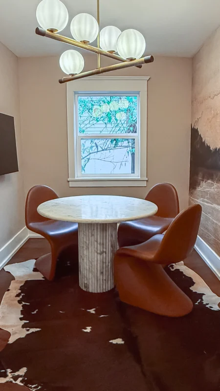 Wide shot of a small meeting room with a round marble table, three brown S-shaped chairs, a cowhide rug, and a modern globular chandelier. A window is in the background.