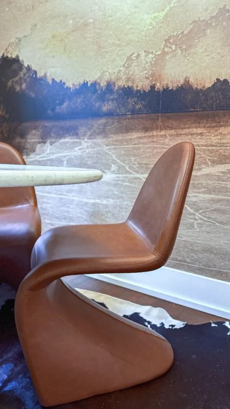 Close-up on the curved back and base of a brown leather-look S-shaped office chair against a textured abstract wall mural.