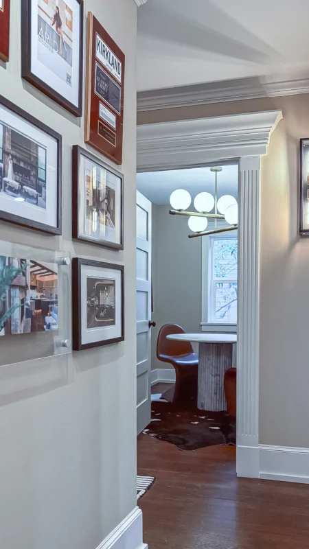 Office hallway with framed artwork and plaques leading into a meeting room with a marble table, brown chair, and a white globular chandelier.