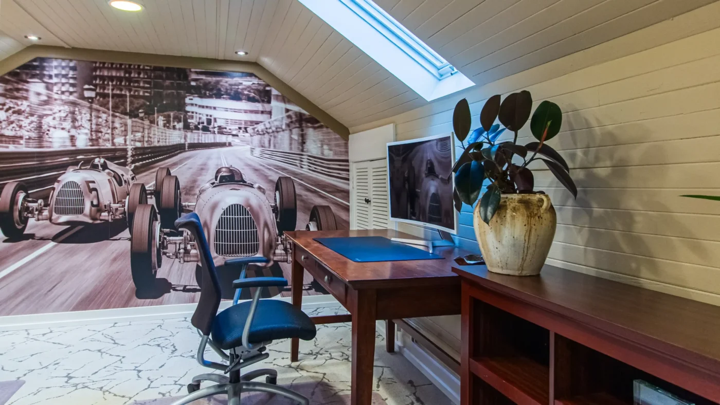 Private attic-style office with a dramatic wall mural of two vintage race cars. A dark wood desk with a computer monitor and a potted rubber plant sits under a ceiling window.