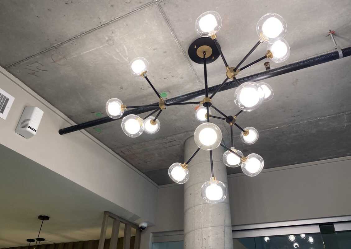 Contemporary multi‑arm black and brass chandelier with round glass shades on exposed concrete ceiling.