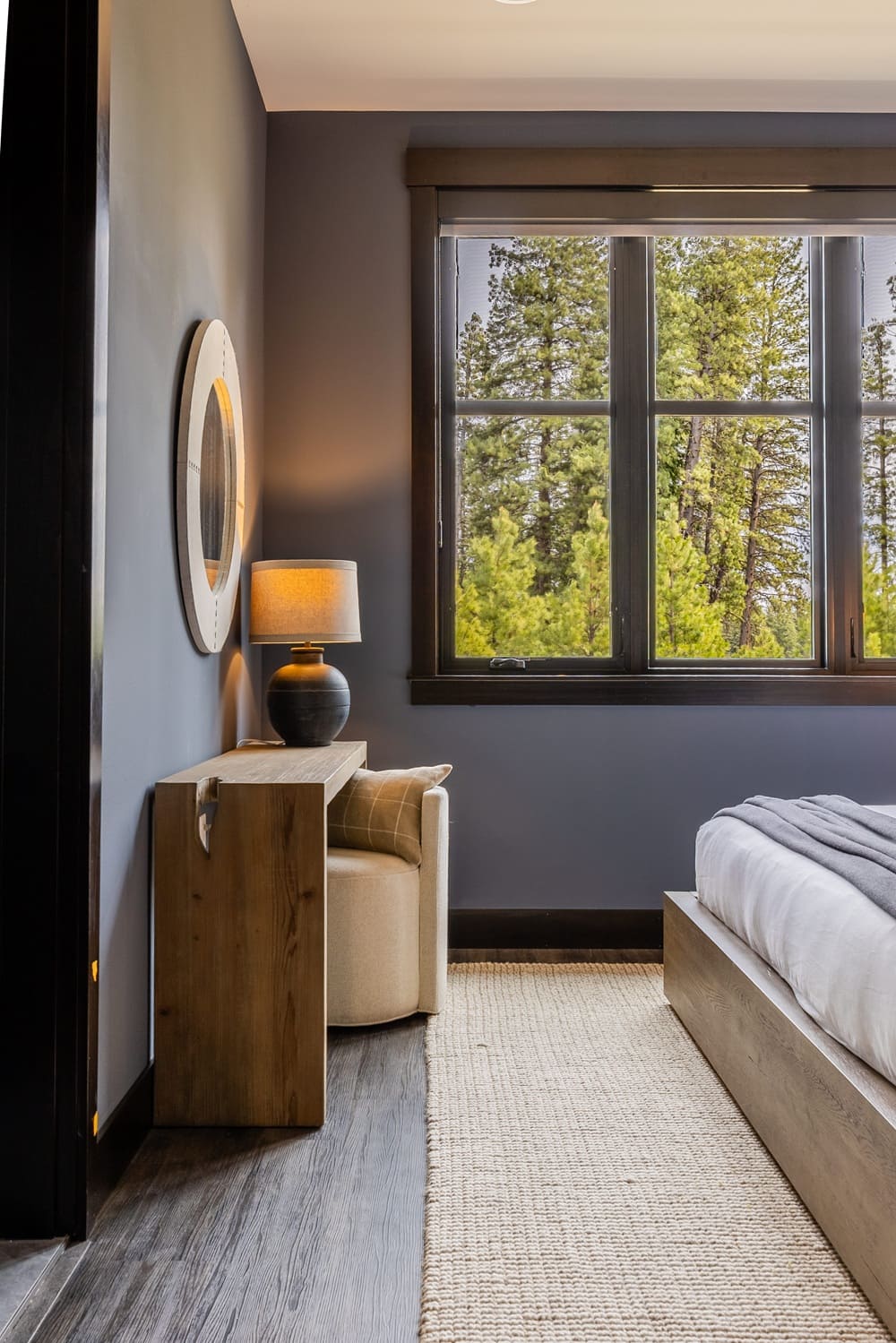 Corner of a blue guest bedroom in the Suncadia home with a wood console table, amber-shade table lamp, round wood-framed mirror, and tall window framing a forest view.