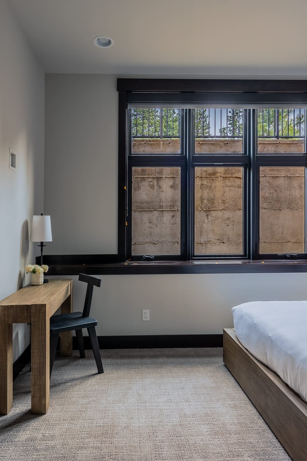 Lower-level guest bedroom in the Suncadia home with a natural wood writing desk, black Windsor-style chair, ceramic table lamp, triple casement window above a wood platform bed, and a textured natural area rug.