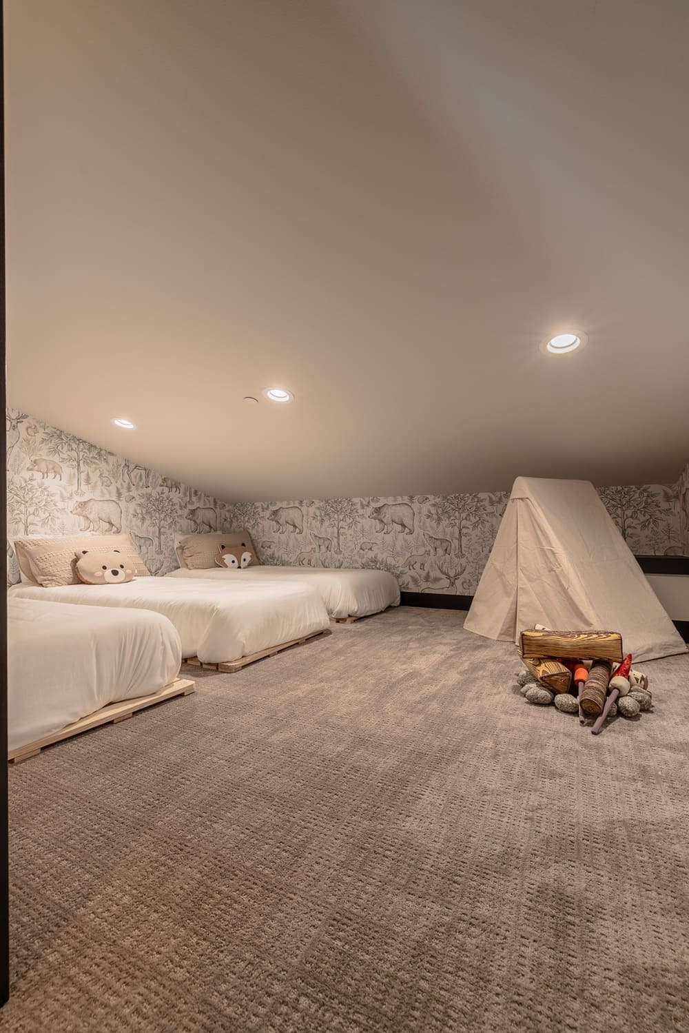 Attic-style kids' sleep nook in the Suncadia home with twin platform beds, forest animal wallpaper, animal plush pillows, a canvas teepee tent, faux campfire setup, and plush wall-to-wall carpet.