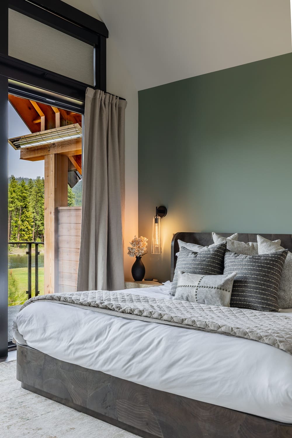 Primary bedroom in the Suncadia home with a sage green accent wall, gray channel-tufted upholstered bed, layered neutral pillows, white quilted throw, brass and glass wall sconce, and a floor-to-ceiling window opening to the forest and lake.
