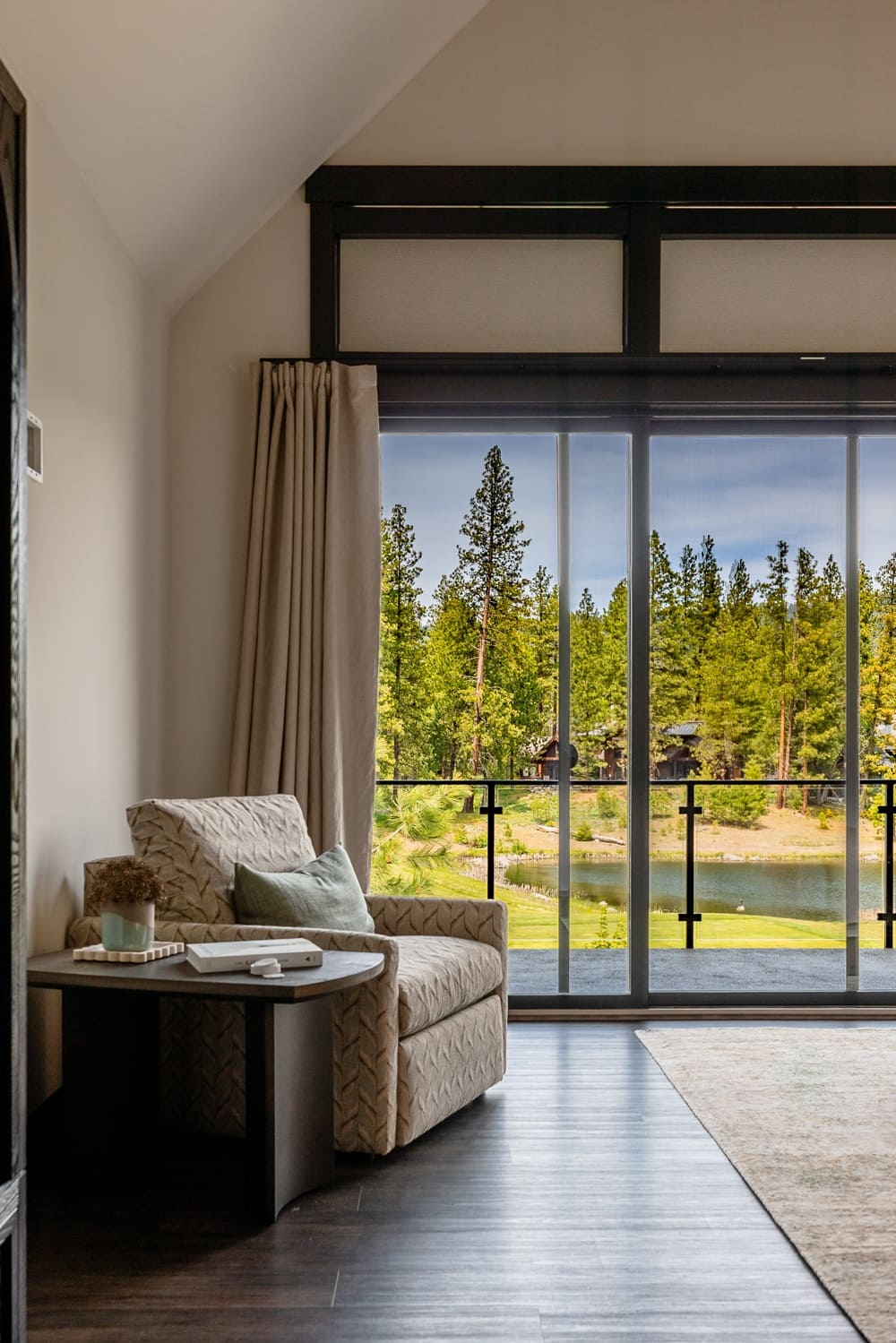 Private reading nook inside a Suncadia bedroom with a textured cream armchair, neutral drapery, round wood side table, and floor-to-ceiling window framing a pond and forest beyond the balcony.