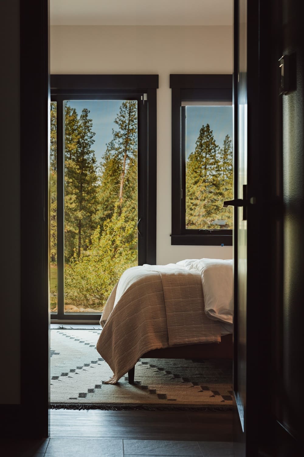 Sunlit view from a hallway into the Suncadia guest bedroom with a camel leather bed, cream quilted throw, kilim patterned area rug, and floor-to-ceiling windows framing pine trees.
