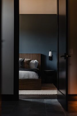 View of the Suncadia blue guest bedroom framed through a dark doorway, showing the wood slat headboard, layered pillows, wood platform bed, black pedestal side table, and wall sconce.