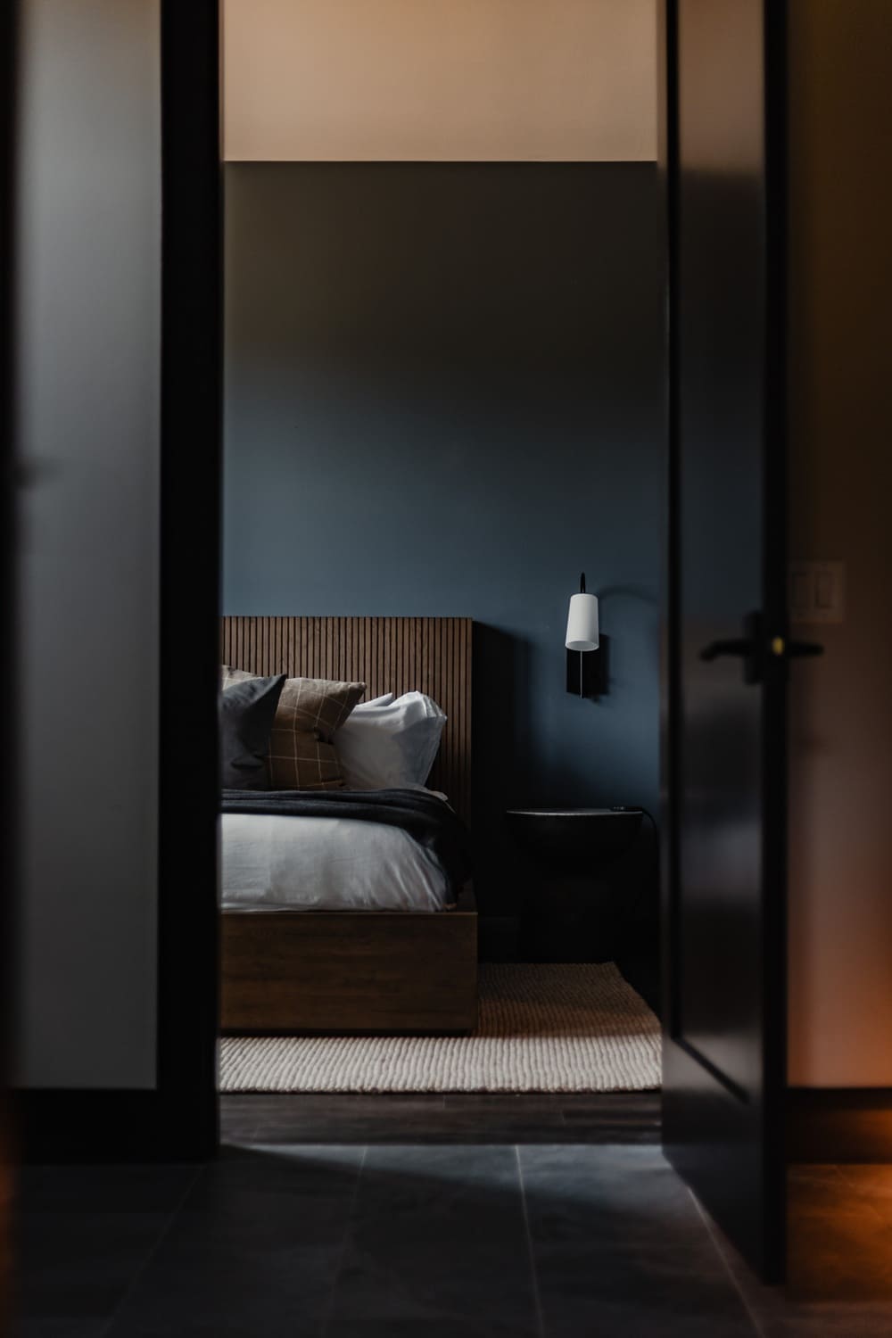 View of the Suncadia blue guest bedroom framed through a dark doorway, showing the wood slat headboard, layered pillows, wood platform bed, black pedestal side table, and wall sconce.