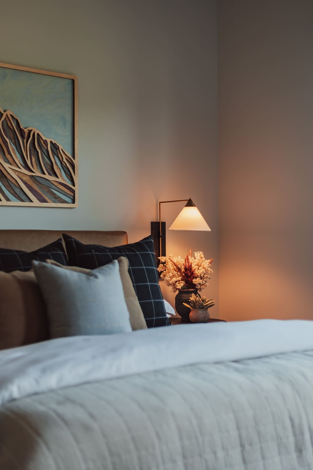 Warmly lit bedside detail in the Suncadia guest bedroom with a camel leather headboard, layered neutral pillows, carved wood mountain art, glowing brass cone sconce, dried florals, and a potted succulent.