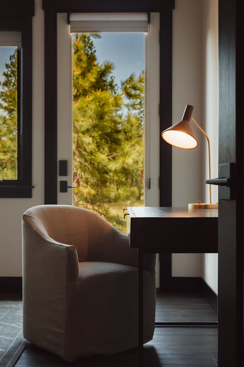 Quiet desk nook inside a Suncadia bedroom with a cream curved armchair, dark stained wood desk, copper cone task lamp, and floor-to-ceiling window framing the surrounding pine forest.