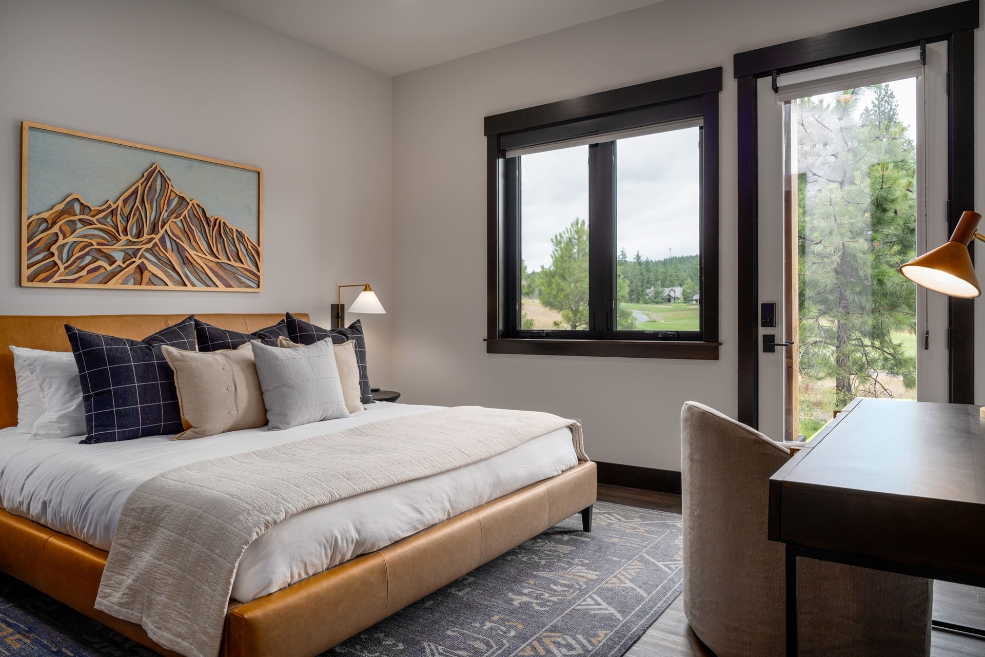Wide view of the Suncadia guest bedroom with a camel leather upholstered bed, 3D carved wood mountain art, brass reading lamp, large window with forest and golf course view, and a writing desk with copper floor lamp.