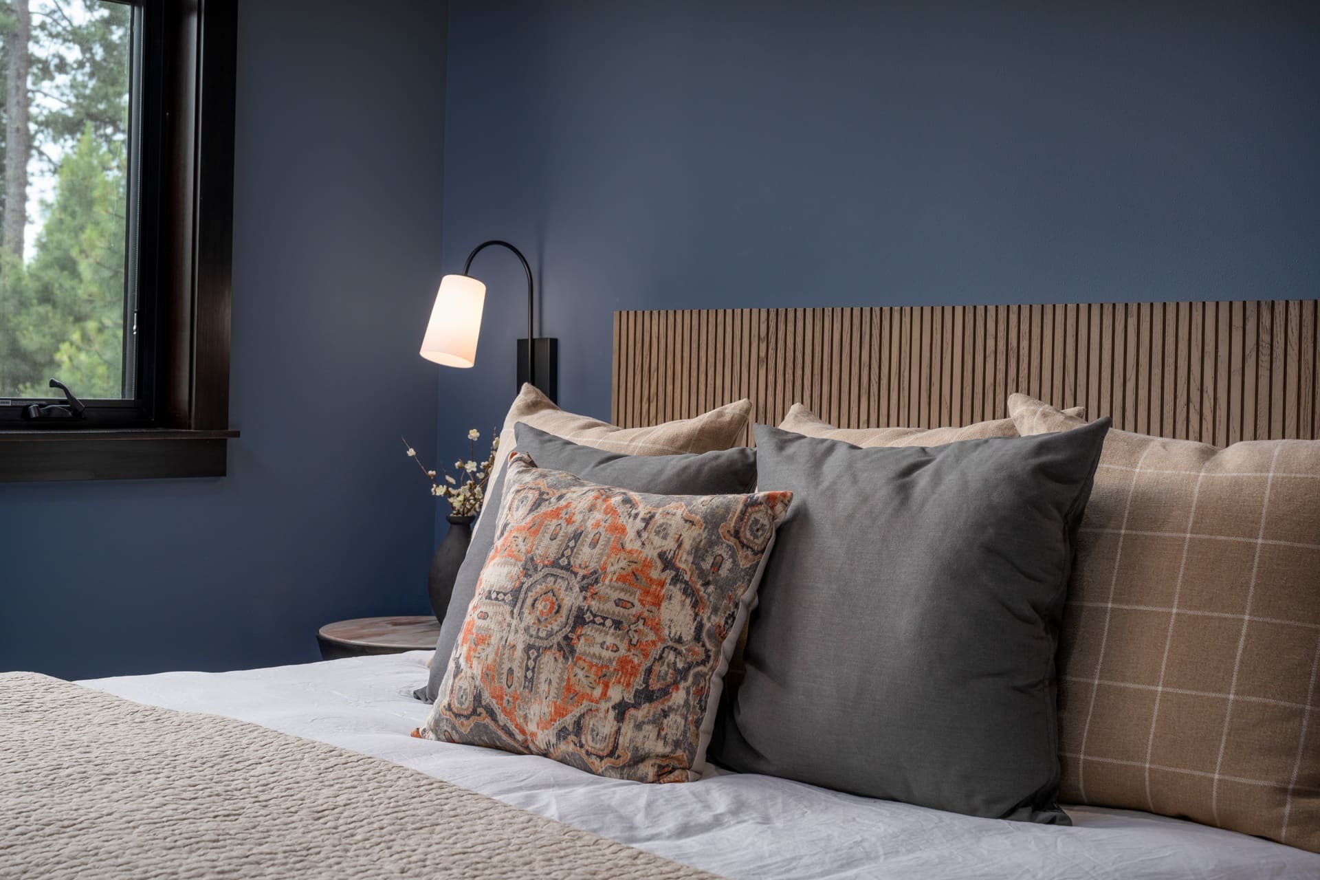 Close-up of layered bedding in the Suncadia blue guest bedroom with a wood slat headboard, charcoal and tan windowpane pillows, a patterned kilim-style accent pillow, and a black reading lamp.