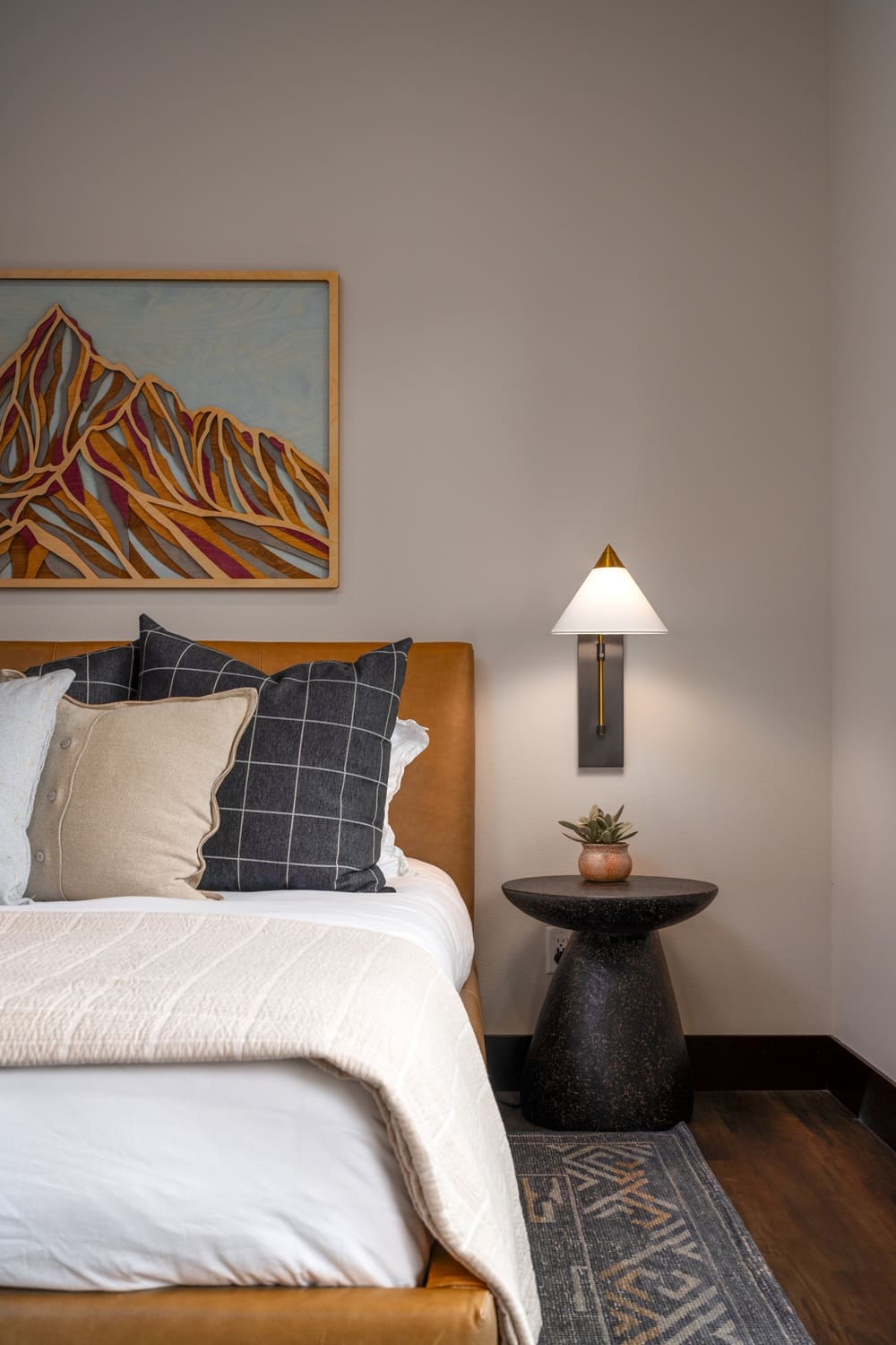 Guest bedroom in the Suncadia home with a camel leather upholstered bed, layered neutral and navy check pillows, a framed mountain landscape painting, brass wall sconce, and a dark pedestal side table.