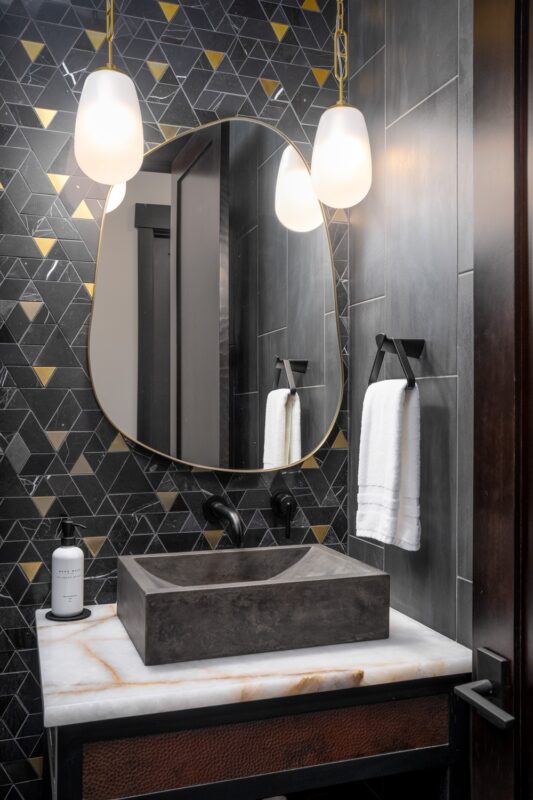 Concrete vessel sink with running water and a matte black wall mount faucet set into a white onyx countertop against black and brass mosaic tile.