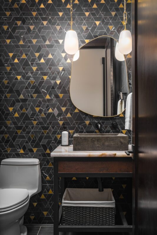 Suncadia powder room featuring a black and brass mosaic feature wall, hammered copper vanity with woven basket storage, and twin brass pendants.