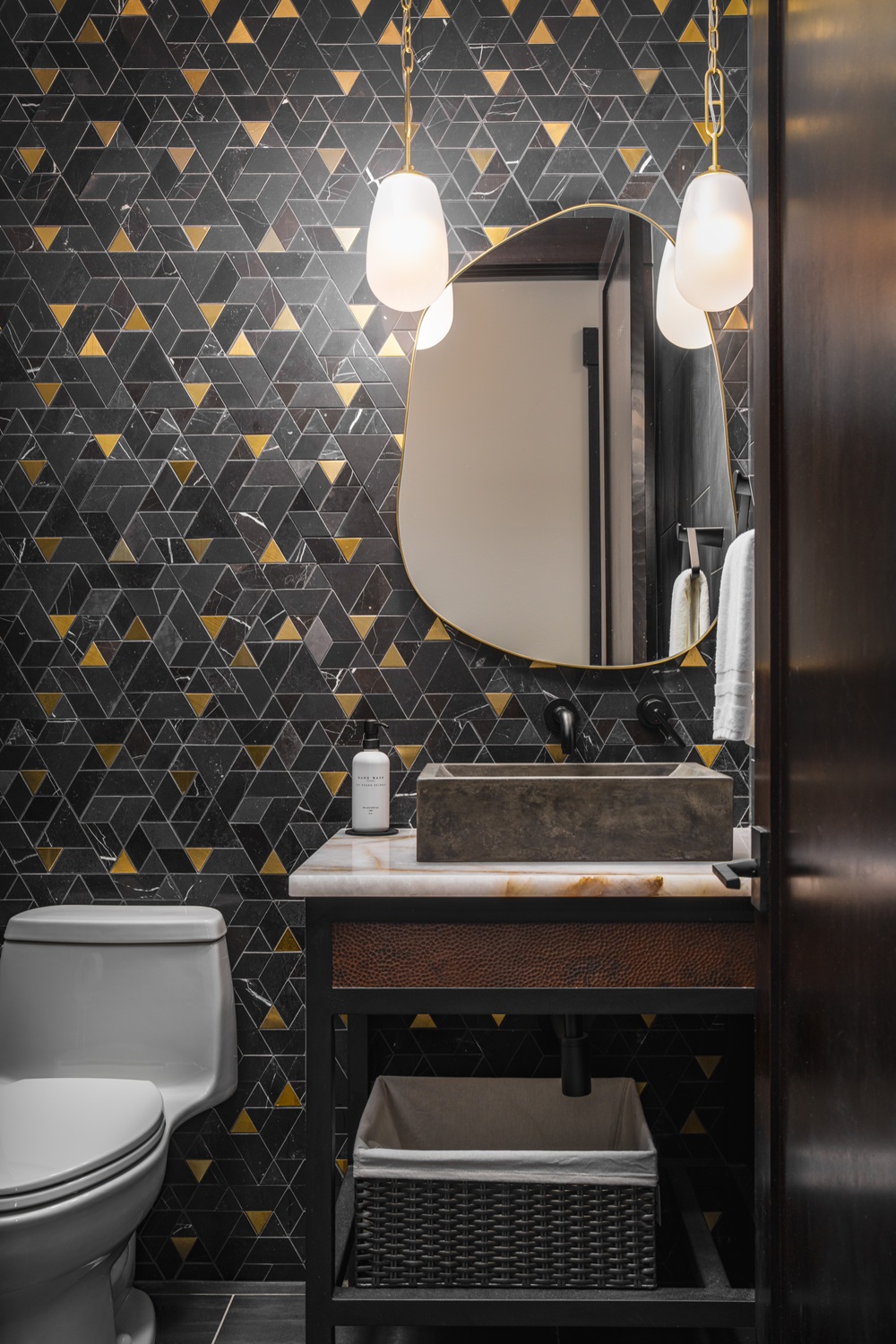Suncadia powder room featuring a black and brass mosaic feature wall, hammered copper vanity with woven basket storage, and twin brass pendants.