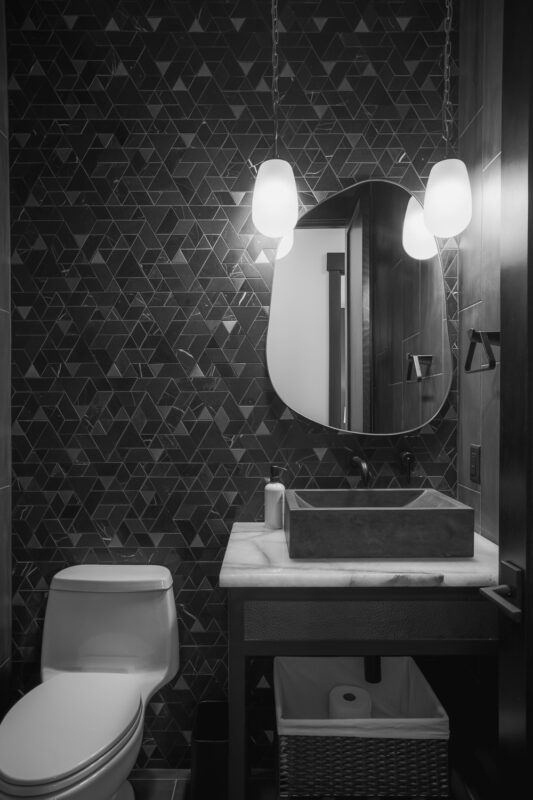 Black and white view of the Suncadia powder room showing the geometric mosaic tile pattern, organic mirror, and dual pendant lights above the vanity.