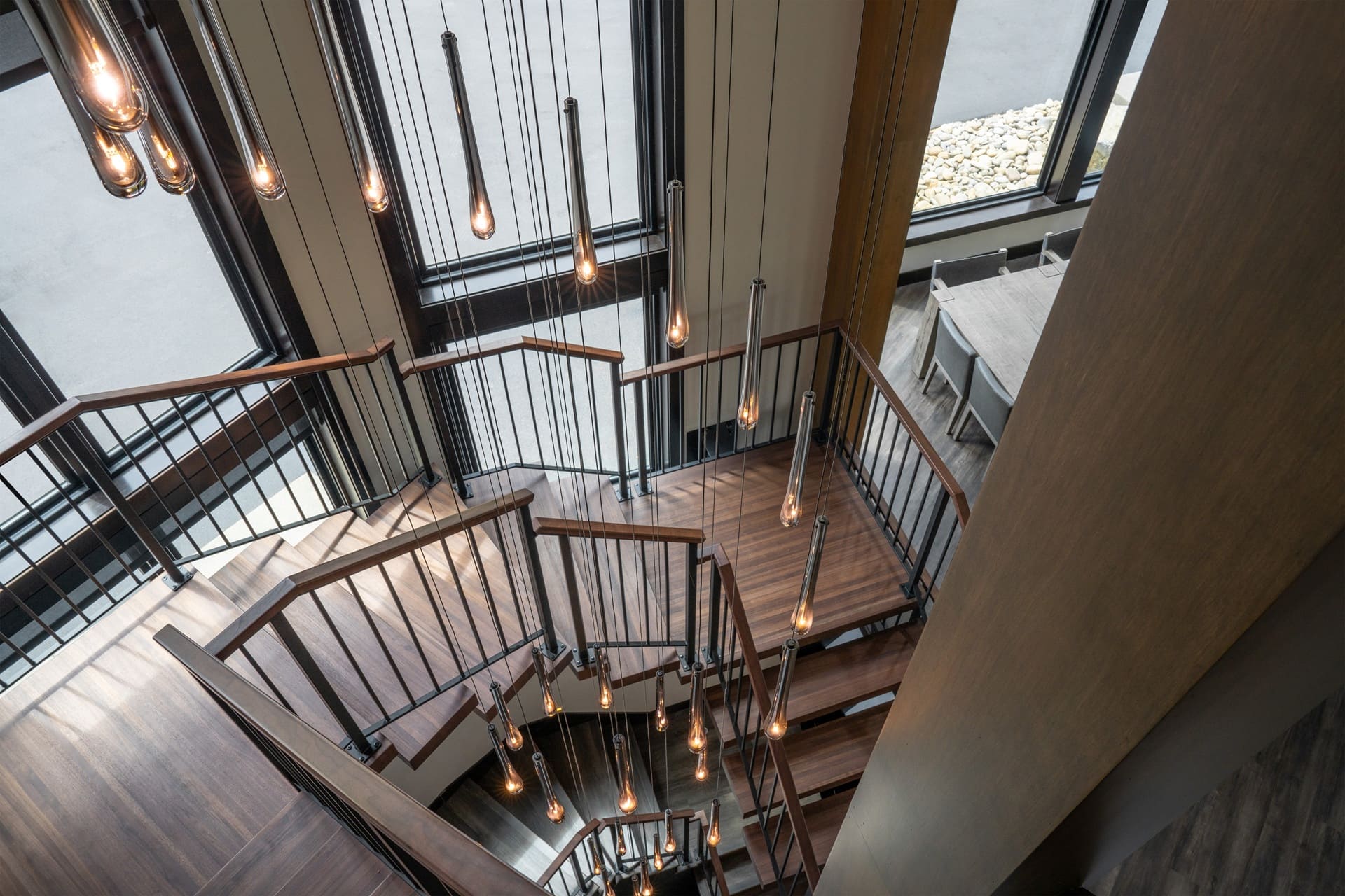 Dramatic overhead view down the Suncadia staircase showing switchback wood treads, black spindle railings, and the full teardrop pendant cascade.