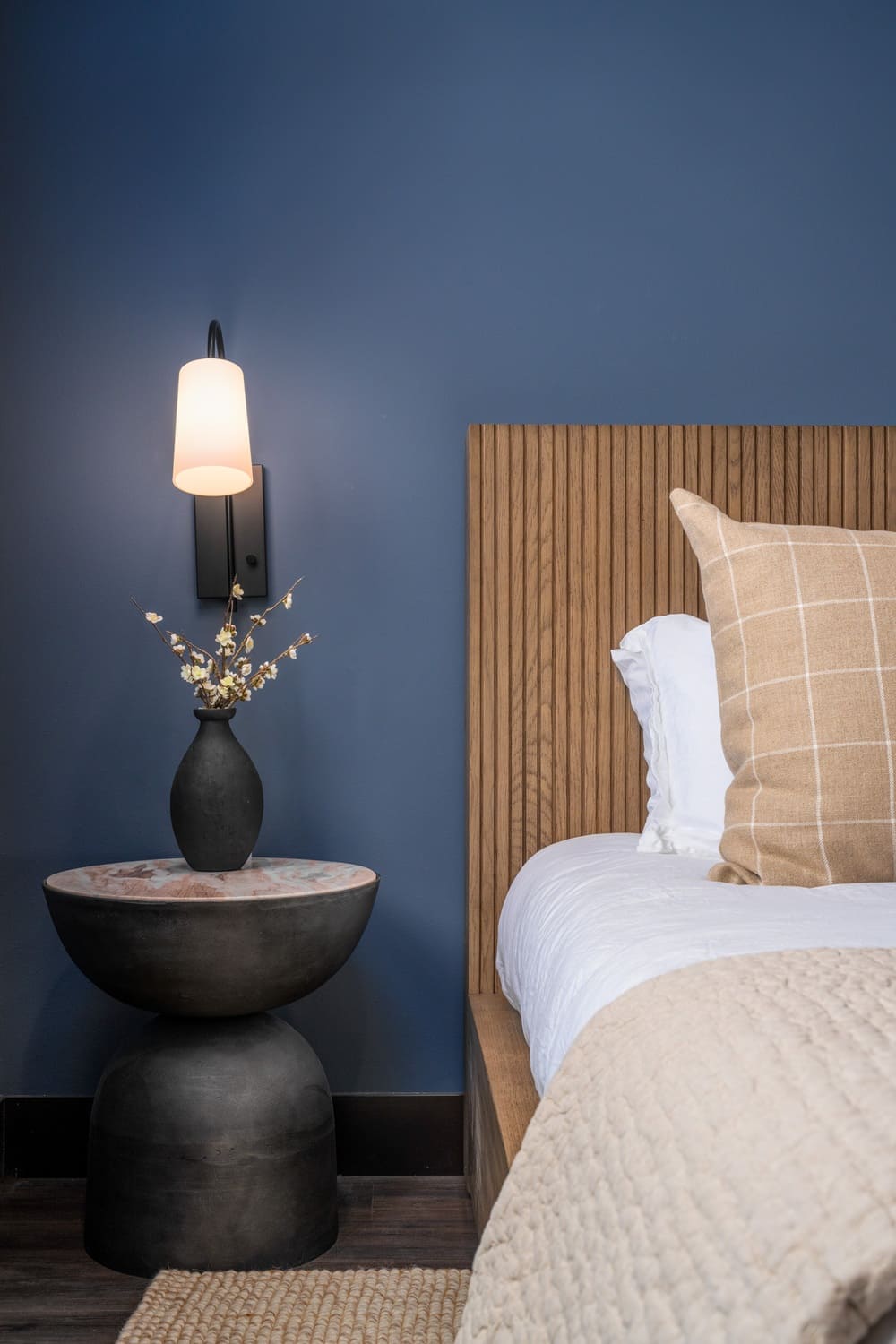 Nightstand vignette in the Suncadia blue guest bedroom with a wood slat headboard, black pedestal side table, ceramic bud vase, and matte black wall sconce with frosted shade.
